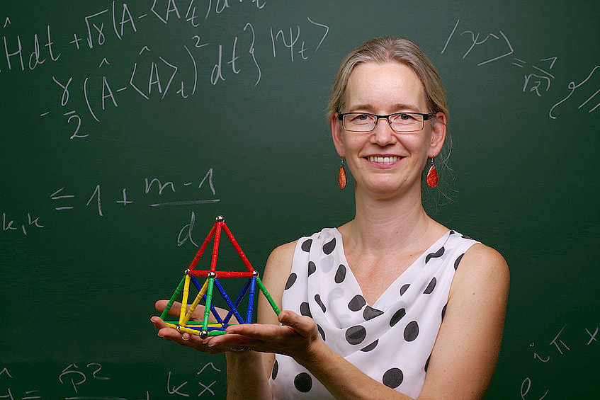 Physik-Uni-Professorin mit Modell aus Streben und vor einer Tafel mit Formeln