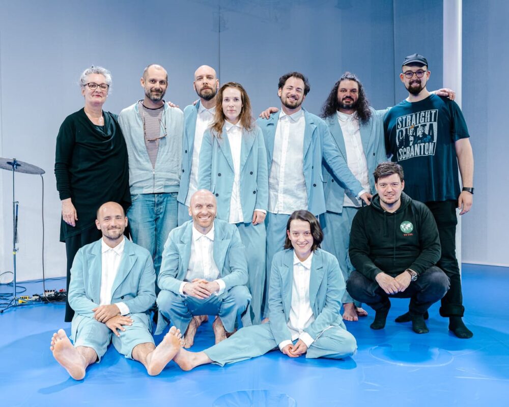 Teamfoto - Schauspieler:innen und Musiker in den blauen Anzügen - sowie Menschen im Hintergrund (Bühne, künstlerische Beratung, Technik)