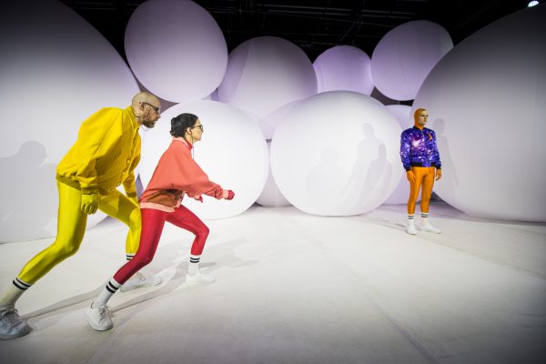 Szenenfoto aus "Rand" - jeweils Schauspieler*innen in bunten, knalligen Sportanzügen vor oder zwischen riesigen weißen, ständig aufgeblasenen Ballons