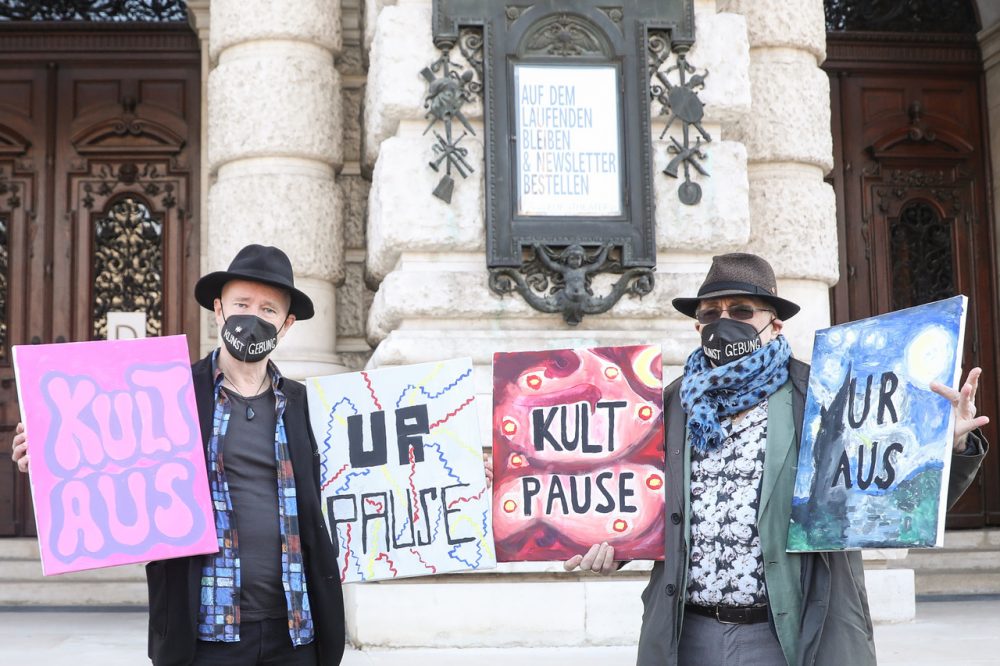 Zwei Männer vor dem geschlossenen Burgtheater. Sie halten Tafeln auf denen sie die Öffnung von Kunst und Kultur als erste nach dem Lockdown fordern