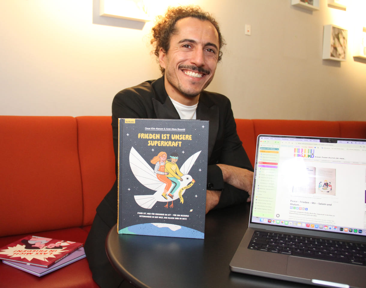 Autor Omar Khir Alanam mit seinem jüngsten Buch "Frieden ist unsere Superkraft" und dem geöffneten Laptop von KiJuKU, wo das Interview mitgeschrieben wurde