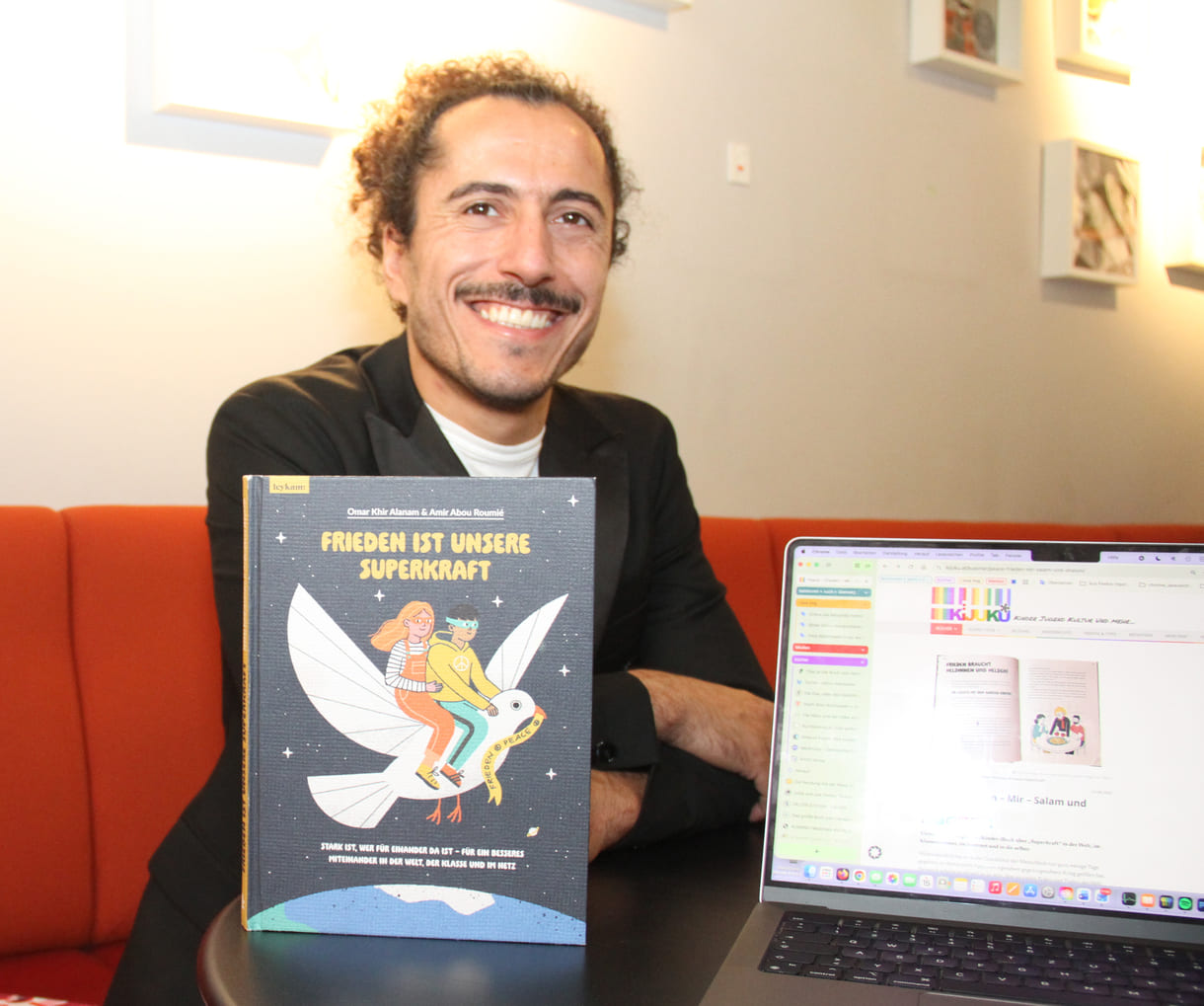 Autor Omar Khir Alanam mit seinem jüngsten Buch "Frieden ist unsere Superkraft" und dem geöffneten Laptop von KiJuKU, wo das Interview mitgeschrieben wurde