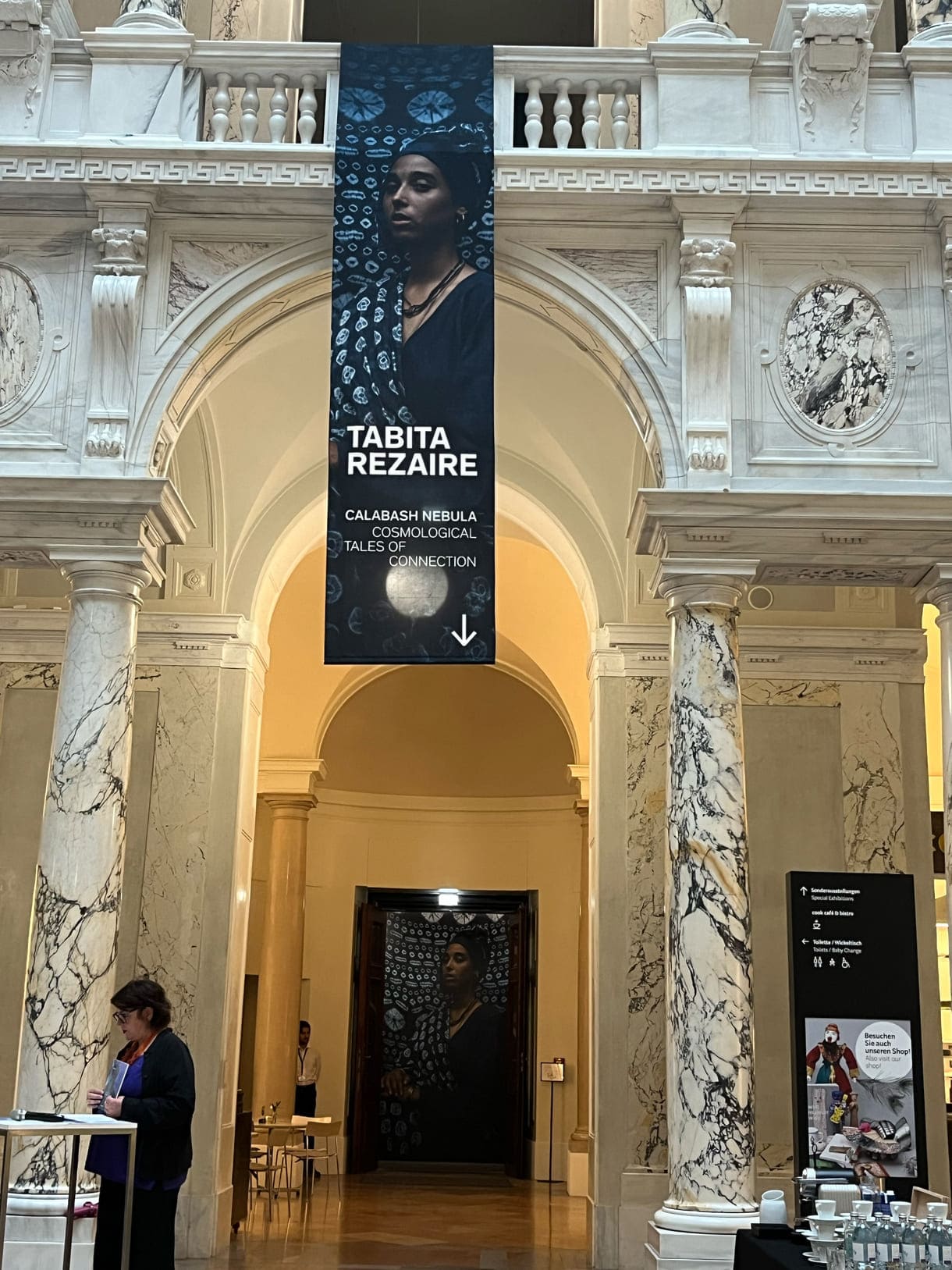 Foto und Banner zur Ausstellung und am Redetisch Claudia Banz, Direktorin des Weltmuseums