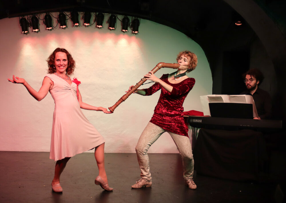 Szenenfoto aus dem musikalischen Kabarettprogramm "Zwa Goschn Oper" - zwei Frauen auf der Bühne, im Hintergrund ein Pianist