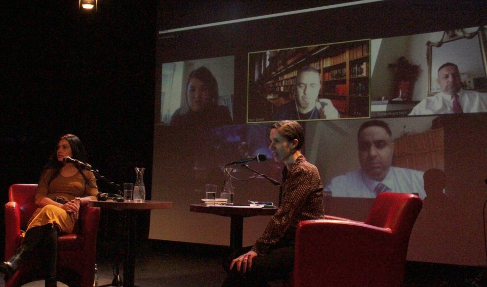 Diskussionsrunde auf der Bühne mit zugeschalteten Online-Gästen auf der Leinwand im Hintergrund