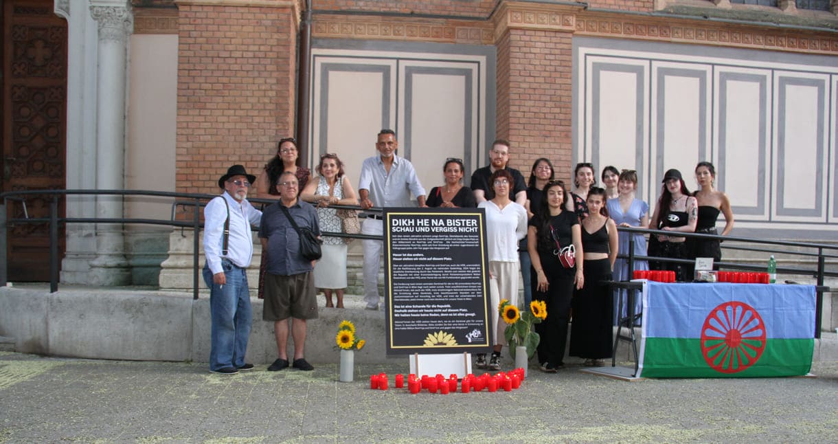 Aktivist:innen und Gedenk-Gäst:innen zwischen Kirche und Plakat sowie Infotisch