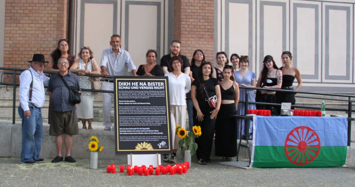 Aktivist:innen und Gedenk-Gäst:innen zwischen Kirche und Plakat sowie Infotisch