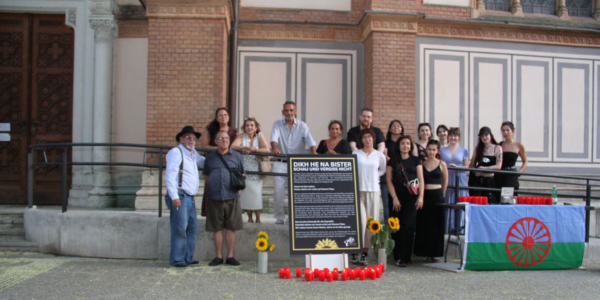 Aktivist:innen und Gedenk-Gäst:innen zwischen Kirche und Plakat sowie Infotisch