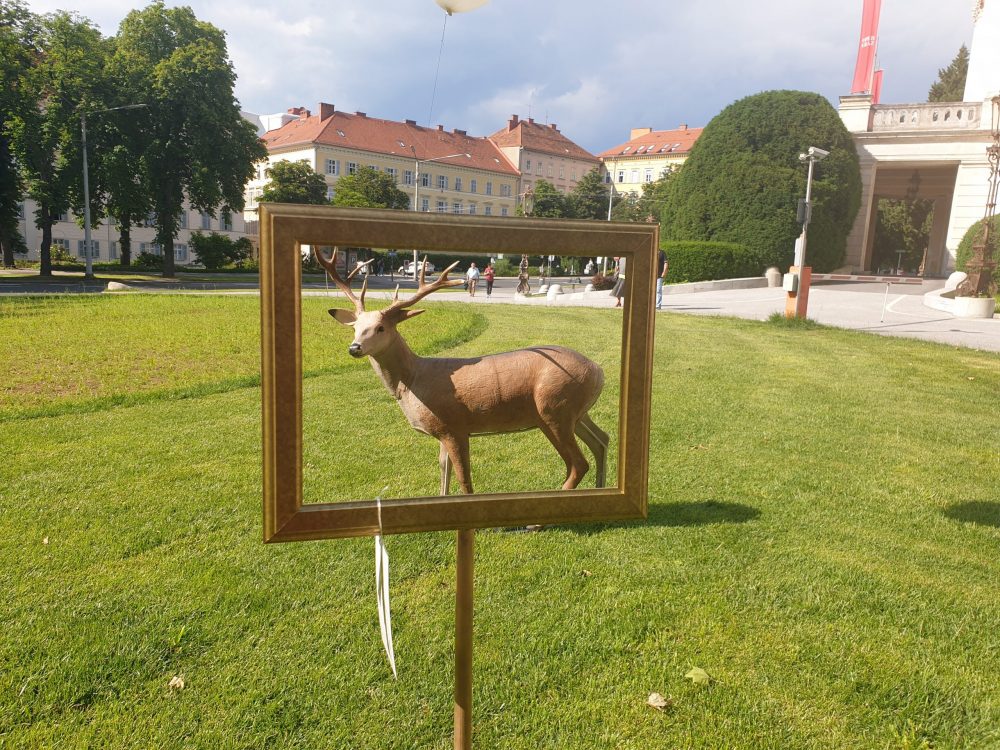 Großer Kunststoff-Hirsch zwischen Grazer Oper und Next Liberty betrachtet durch einen leeren Bilderrahmen