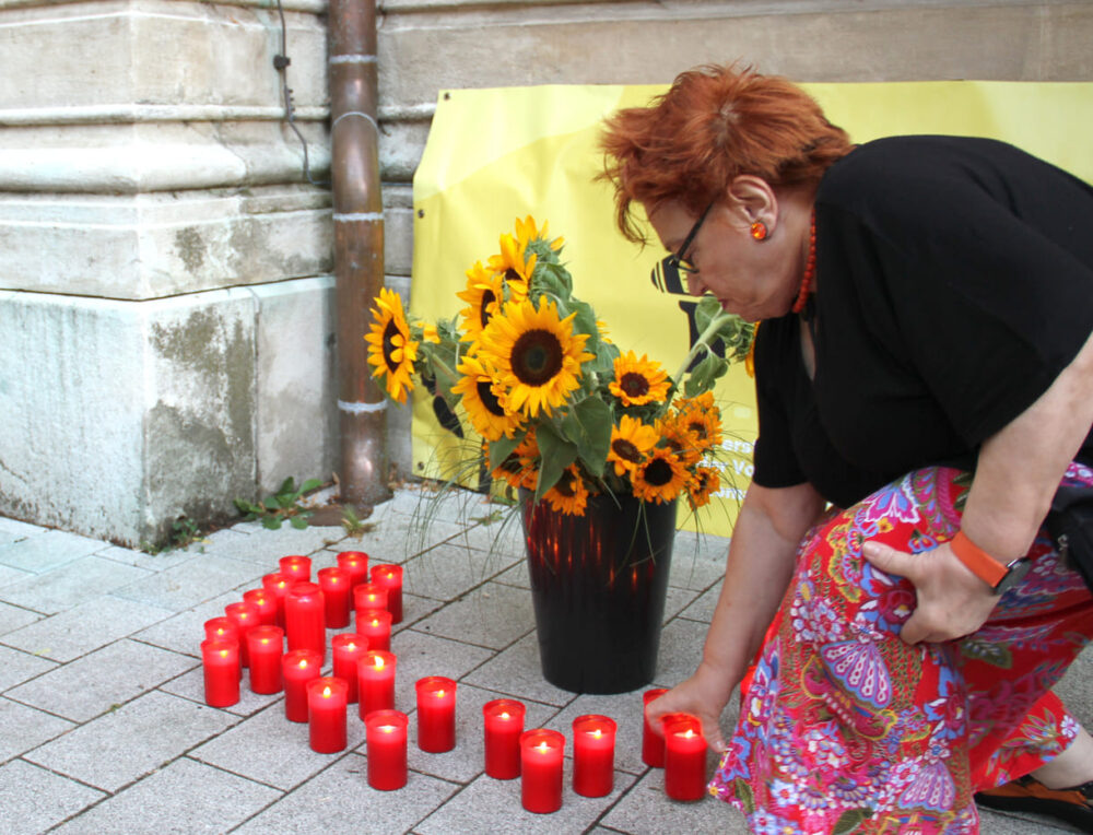 Teilnehmende stellen ihre Kerzen zu den Sonnenblumen und dem Transparent der Hör, der Hochschüler*innenschaft Österreichischer Roma und Romnja