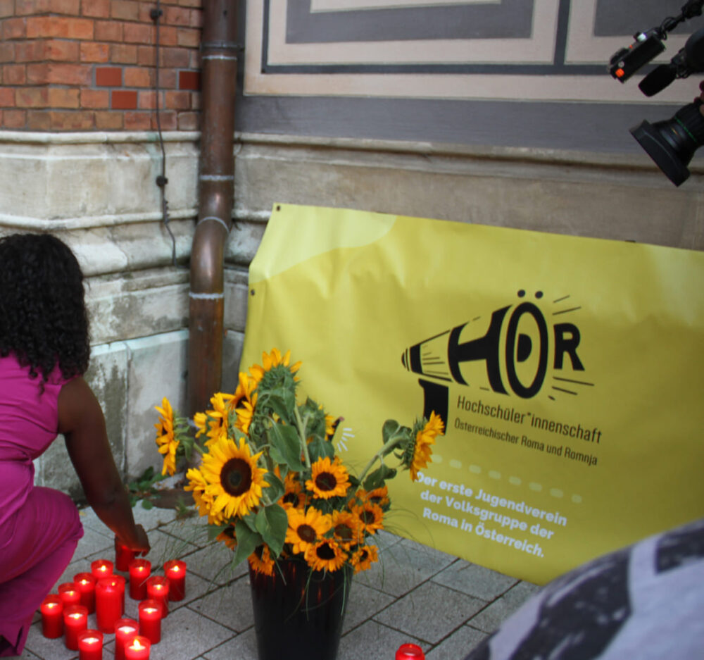 Teilnehmende stellen ihre Kerzen zu den Sonnenblumen und dem Transparent der Hör, der Hochschüler*innenschaft Österreichischer Roma und Romnja