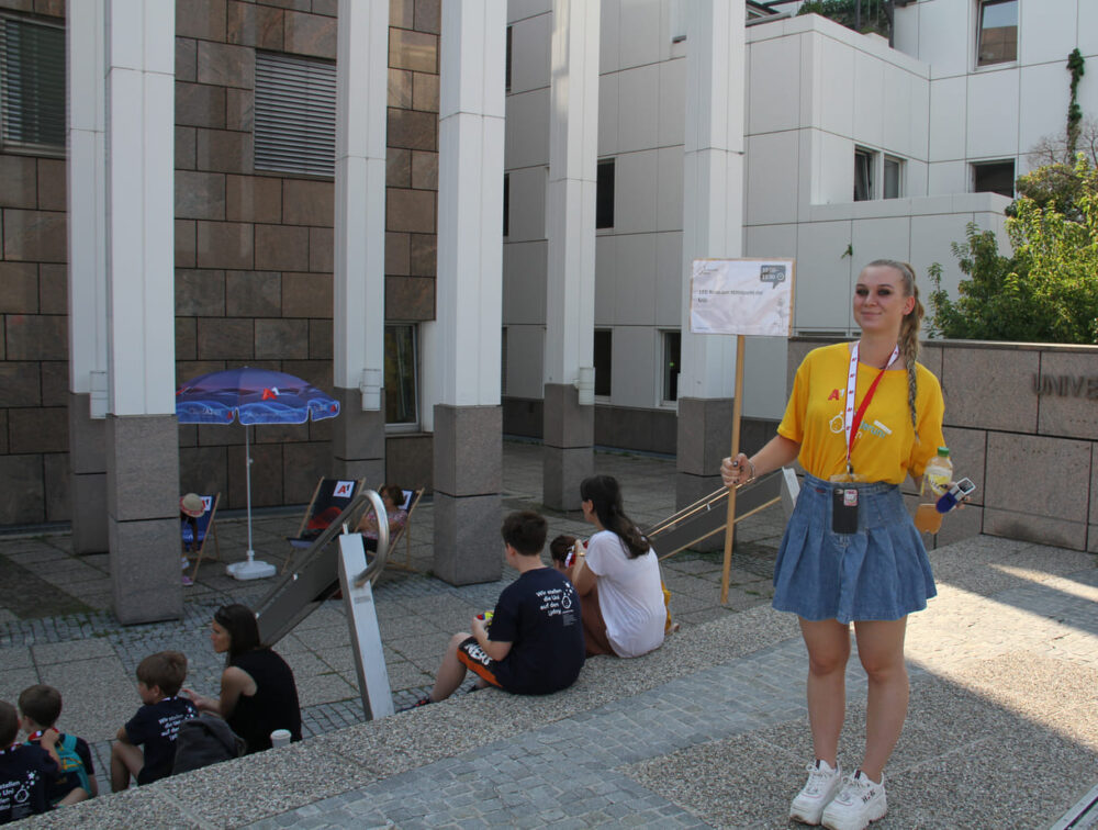 Volunteers in gelben Kinderuni-T-Shirts halten die Schilder mit den Titeln der Lehrveranstaltungen udn begleiten die Jung- und Jüngststudierenden zu ihren Hörsälen