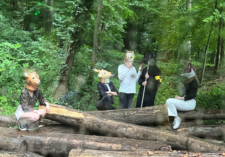 Jugendliche Schauspieler:innen mit Tiermasken kommen aus dem Wald auf den Tour-Bus zu...