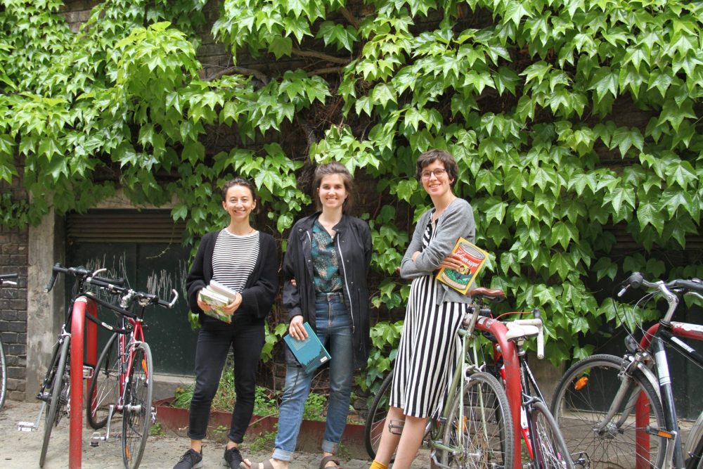Drei junge Frauen mit Büchern stehen im WuK-Hof vor einer begrünten Wand und zwischen Fahrrädern: Kathi Pech, Sara Schausberger und Greta Egle