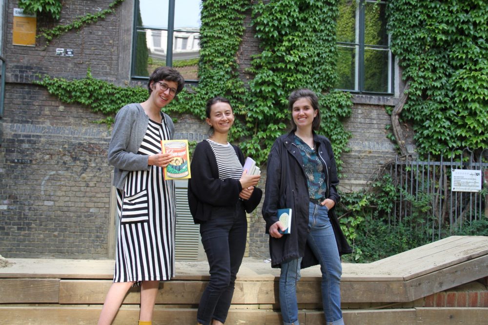 Drei junge Frauen mit Büchern stehen vor einer Holzumrandung der Sandksite und einer begrünten Wand im WuK-Hof: Greta Egle, Kathi Pech und Sara Schausberger