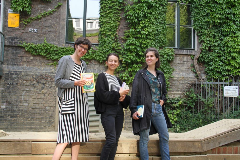 Drei junge Frauen mit Büchern stehen vor einer Holzumrandung der Sandksite und einer begrünten Wand im WuK-Hof: Greta Egle, Kathi Pech und Sara Schausberger