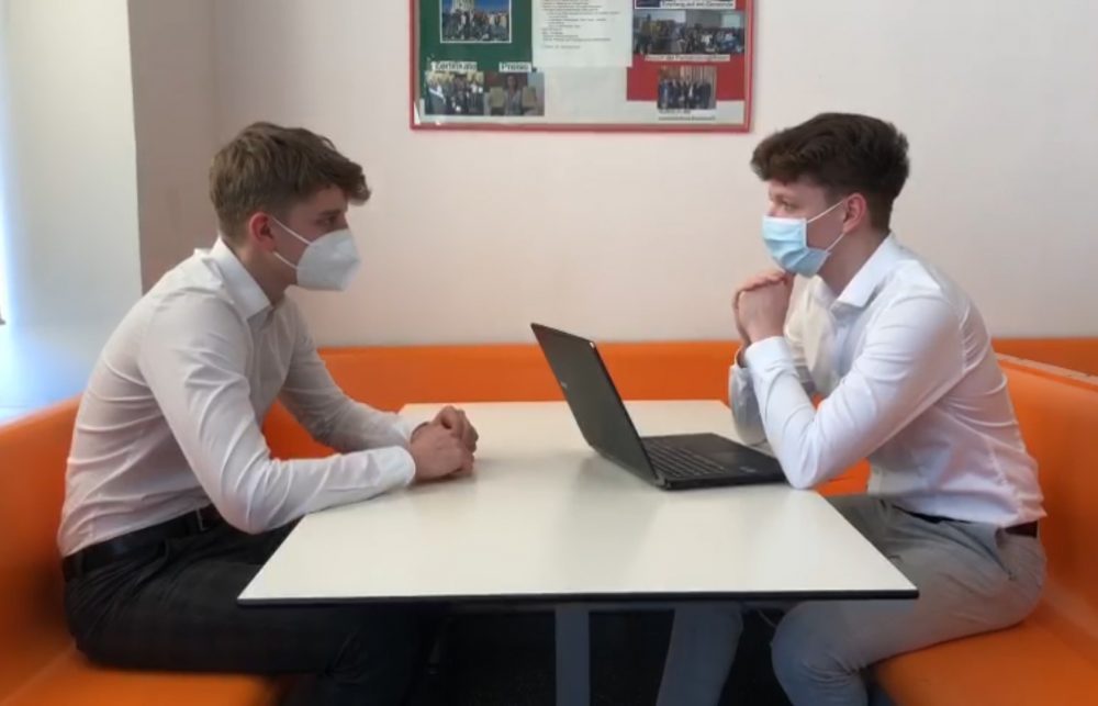 Zwei Jugendliche sitzen einander gegenüber, einer mit Laptop, beide mit FFP2-Masken