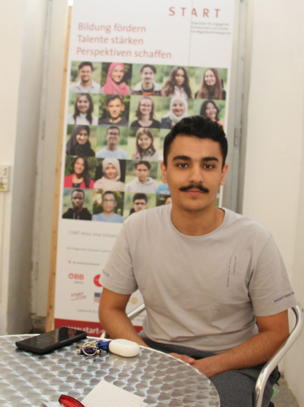 Der junge Fotograf Omran Almasri vor einem Roll-Up des Projekts Start-Stipendium
