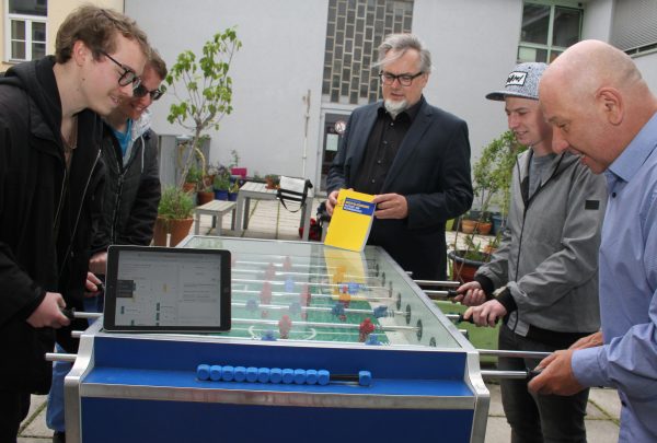 Schüler und Lehrer spielen Tischfußball, ein Laptop mit der Schulhomepage und der Direktor mit Buch zur Digitalisierung